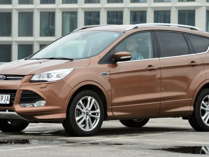 Brązowy Ford Kuga 2019 z kierowcą w okularach, zaparkowany na mokrym parkingu przed nowoczesnym budynkiem.