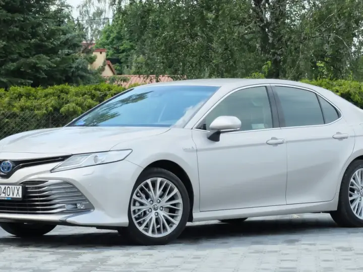 Srebrna Toyota Camry 2.5 Hybrid stoi na wybrukowanym podjeździe, otoczona zielenią.