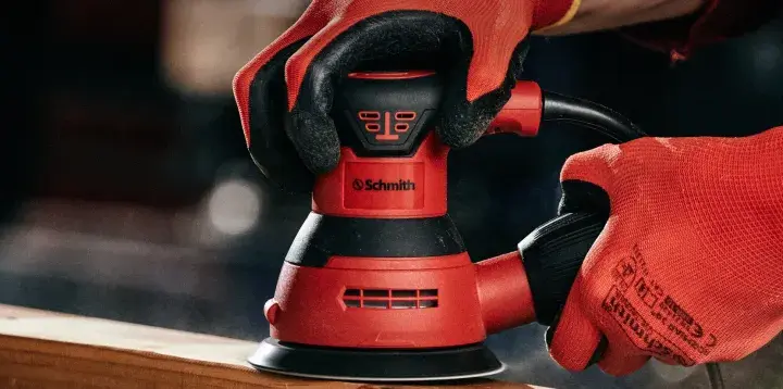 Dłonie w czerwonych rękawiczkach trzymają okrągłą szlifierkę oscylacyjną, gotową do pracy. Różne rodzaje szlifierek pomagają w obróbce drewna.