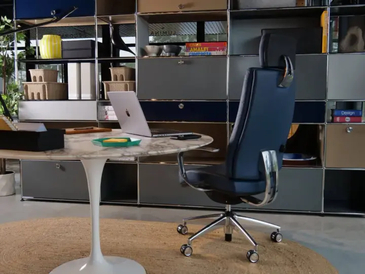 Un fauteuil de bureau design bleu marine, un bureau rond en marbre et un ordinateur portable créent un espace de travail élégant.