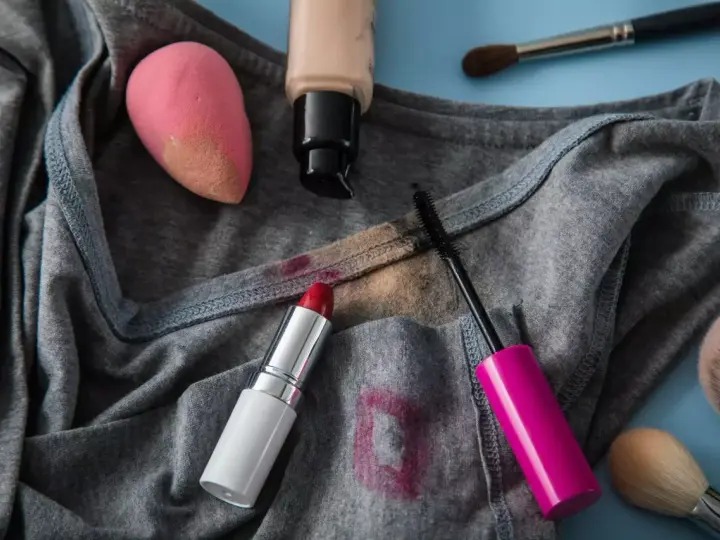 Lippenstiftflecken entfernen: Roter Lippenstift, Mascara, Foundation und Make-up-Schwamm auf grauer Kleidung.