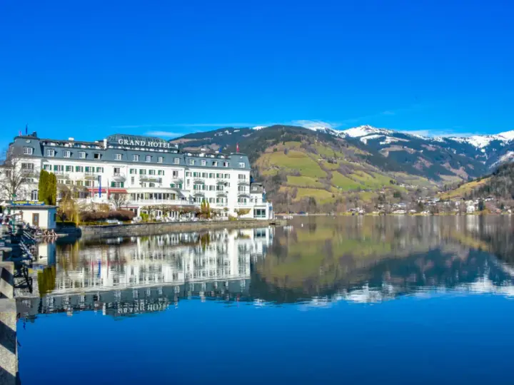 Grand Hotel nad jeziorem w Zell am See, z odbiciem w spokojnej wodzie i ośnieżonymi górami w tle.