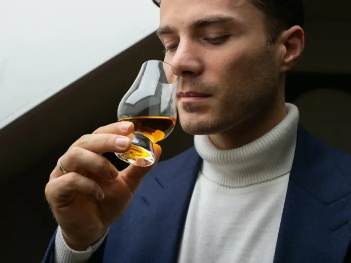 Mężczyzna w eleganckim garniturze i golfie wącha bursztynowy płyn w kieliszku. Czy to single malt?