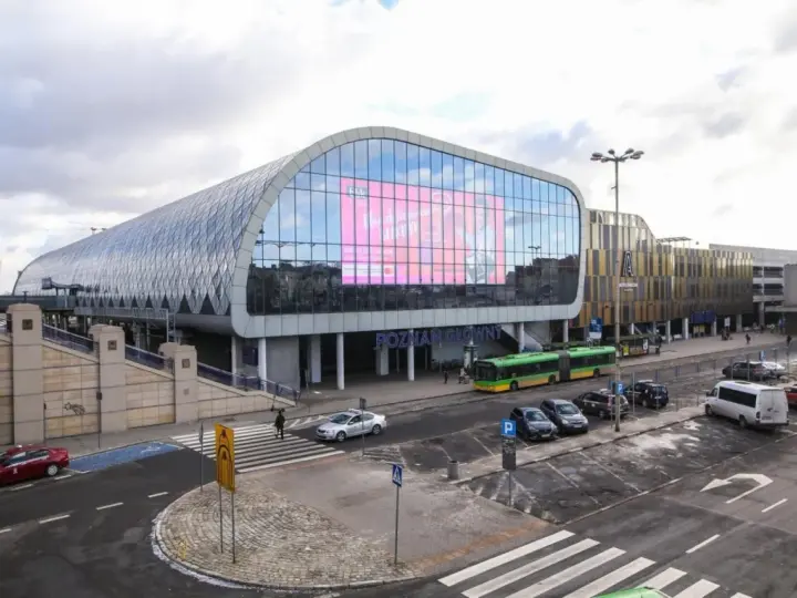 Nowoczesny Poznań Główny z charakterystyczną szklaną fasadą i ekranem. Autobusy i samochody czekają na pasażerów.