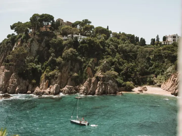 A sailboat floats in turquoise water near a rocky, tree-covered cliff with buildings. Perfect for weekend trips from Barcelona by train.