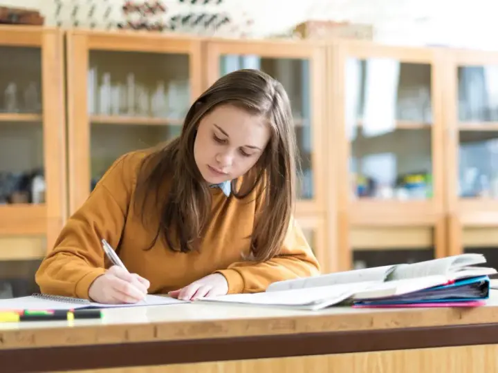 Dziewczyna pisze w zeszycie, przygotowując się do egzaminu. Marzy o stypendium szkolnym, które pomoże jej w dalszej nauce.