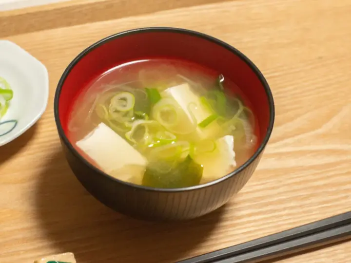 Un bol de soupe japonaise, avec du tofu et des oignons verts, servi avec des baguettes sur un plateau en bois.