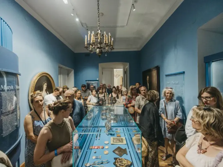 Grupa zwiedzających ogląda wystawę w muzeum. Wśród eksponatów w gablocie widać ozdobne przedmioty, a na ścianach obrazy i tablice informacyjne. Klasyczny wystrój sali z żyrandolem stanowi tło dla tego współczesnego zainteresowania historią.