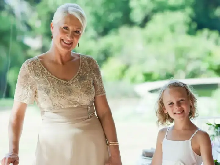 Elegancka stylizacja na komunię dla babci. Uśmiechnięta kobieta w beżowej sukni z haftem i dziewczynka w bieli.