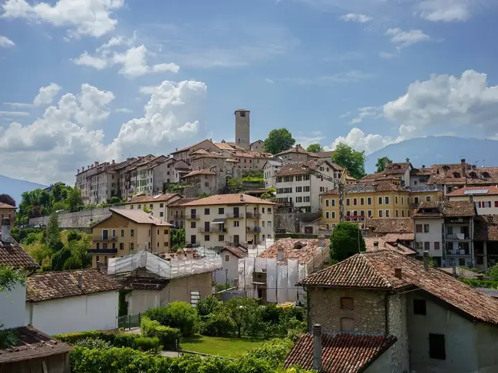 Panoramica di Feltre e dintorni, con case dai tetti rossi che si arrampicano sulla collina sotto un cielo azzurro e nuvoloso.