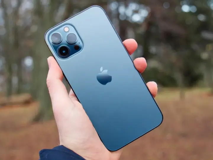 En hand håller en blå iPhone 12 Pro Max. Dess storlek är perfekt för att fånga höstens färger i bakgrunden.