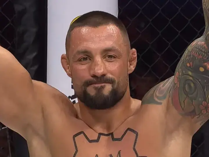 Mateusz Janur, wojownik MMA, z podniesionymi rękami i tatuażami na ramionach i klatce piersiowej, świętuje zwycięstwo.