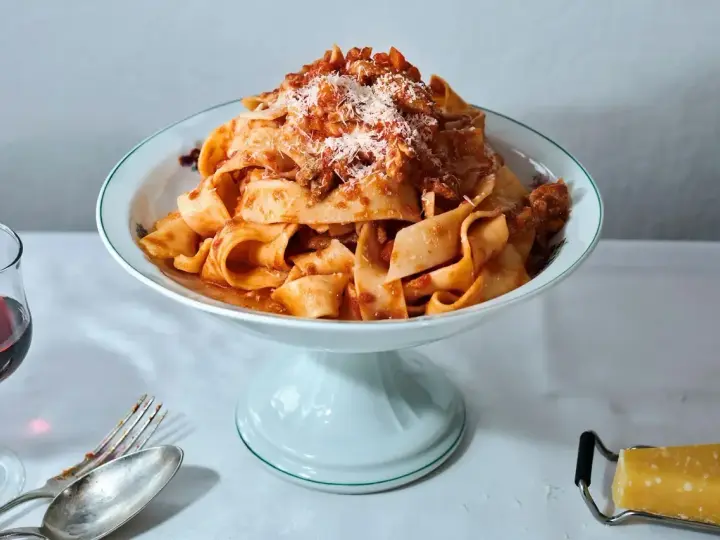 Ein Teller breiter Bandnudeln mit herzhaftem al ragu, bestreut mit Parmesan. Daneben ein Glas Rotwein und Besteck.