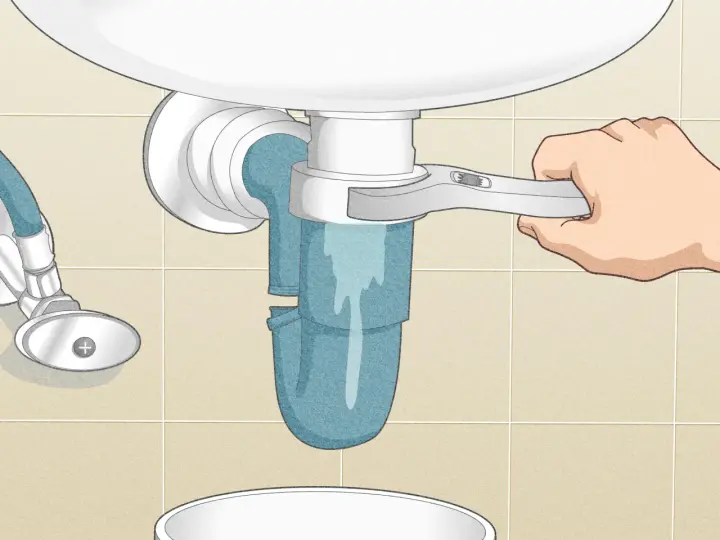 A hand uses a wrench to loosen a sink trap, a step in how to clean a sink drain. Water drains into a bucket below.