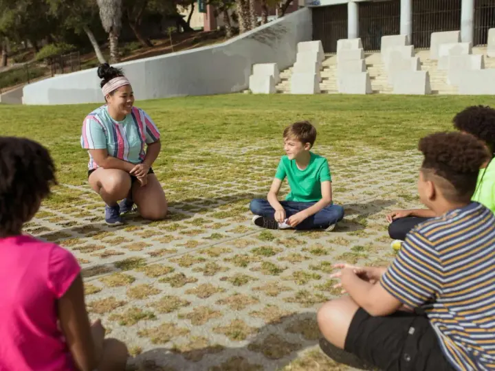 Une animatrice discute avec des enfants assis en cercle dans un parc.