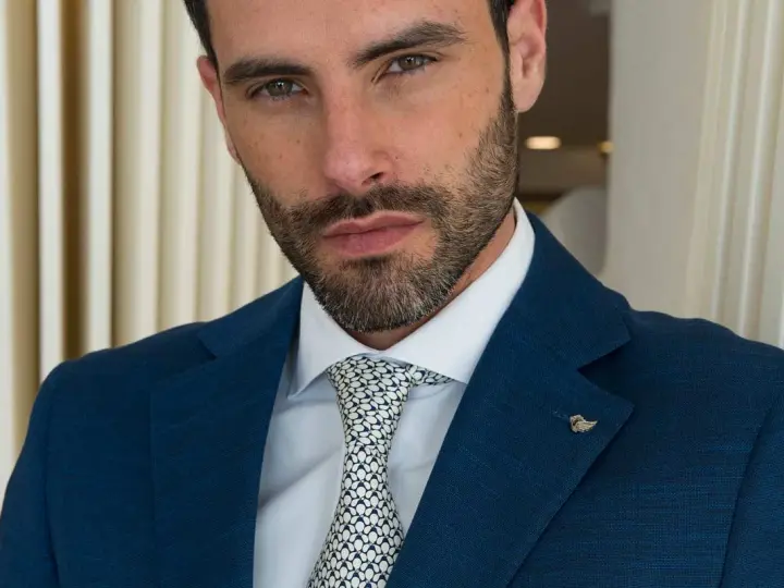 Uomo con abito blu, camicia bianca e cravatta con fantasia a pois bianchi e neri. Un tocco di stile con un pin a forma di ala.