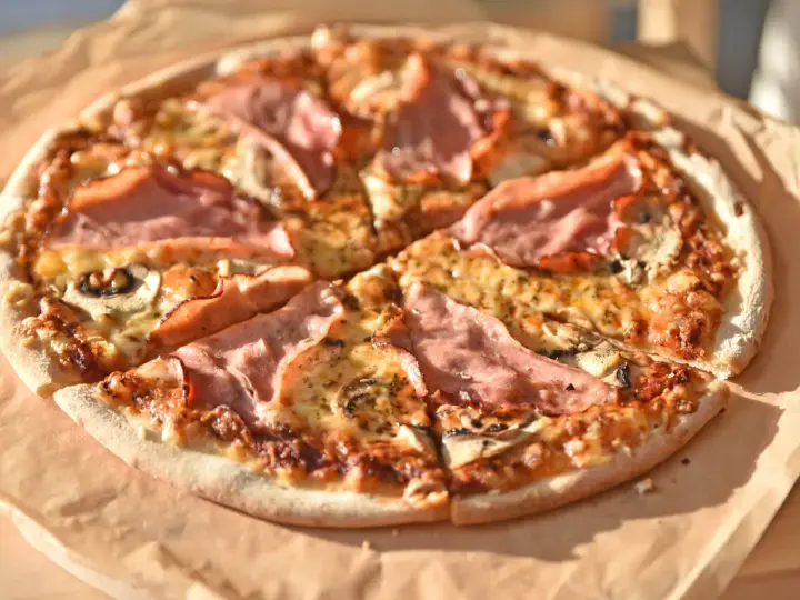 Pizza z szynką i pieczarkami, idealna na szybki obiad. Jakie pieczarki do pizzy? Te ciemne, pokrojone w plasterki, świetnie komponują się z serem i wędliną.