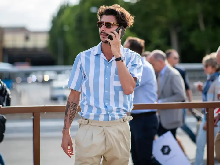 Hombre con camisa a rayas azules y blancas y tiro pantalón alto color beige, hablando por teléfono.