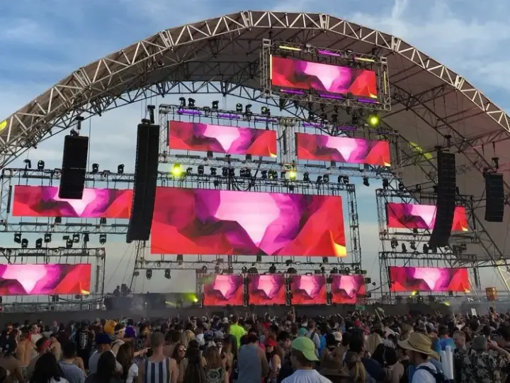 Scène de festival avec écrans LED diffusant des visuels abstraits roses et violets. Une foule danse sous la structure, attendant leur artiste deep house préféré.