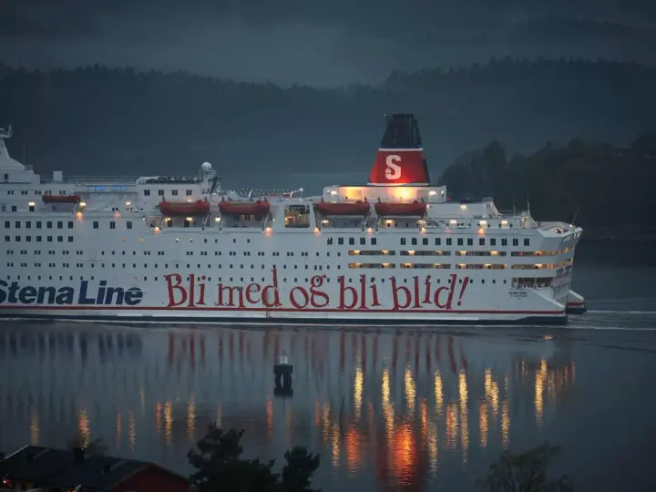 Duży biały prom Stena Line z czerwonym napisem "Bli med og bli blid!" płynie po wodzie o zmierzchu.