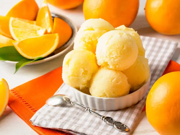 Erfrischendes orangen sorbet in einer Schale, umgeben von frischen Orangen und saftigen Orangenstücken. Ein Löffel liegt bereit.