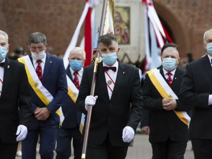 Rycerze Kolumba w garniturach i maskach maszerują z flagą.