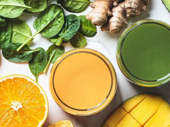 Färgglada juicer, en med apelsin och en grön, omgivna av färska ingredienser som spenat, ingefära, mango och avokado. Perfekt för en hälsosam "te de curcuma y jengibre"-inspirerad smoothie.