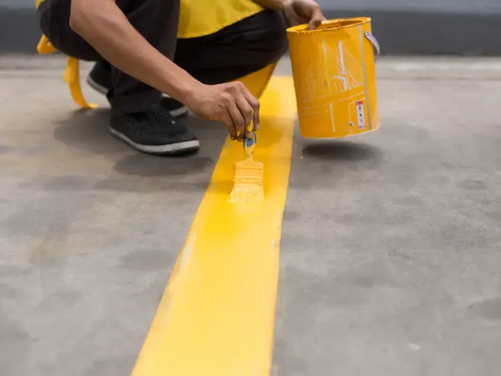 Persona pintando una línea amarilla brillante sobre un suelo de cemento con una brocha.