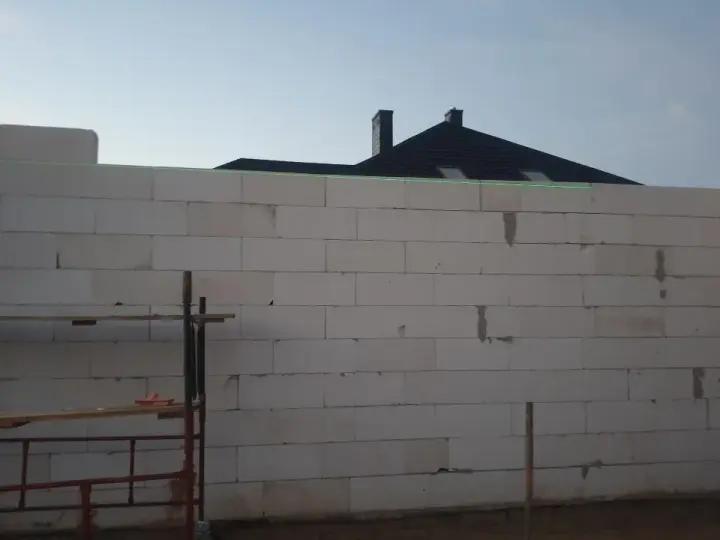 Budowa domu w toku. Ściany z bloczków betonowych czekają na tynkowanie. W tle widać dach i kominy.