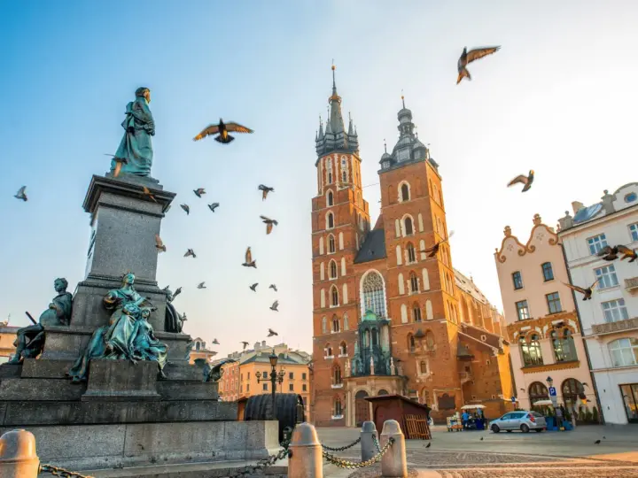 A monument and St. Mary's Basilica dominate this Krakow travel scene. Birds fly around the historic architecture under a clear sky.