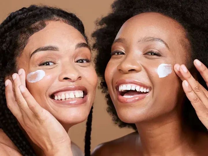 Deux femmes souriantes appliquent une crème sur leur visage, un geste pour une exfoliation visage réussie.