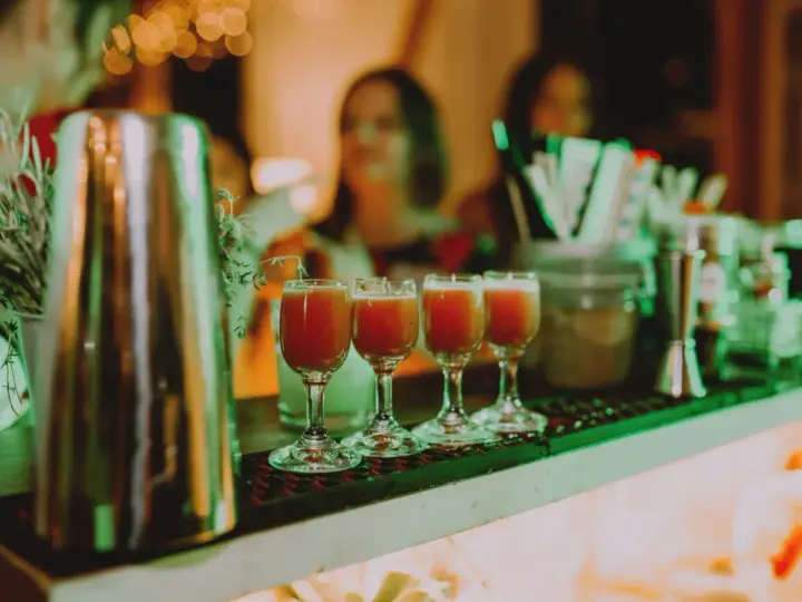 Cztery kieliszki z czerwonym drinkiem stoją na barze, gotowe do podania na weselu. W tle widać gości i dekoracje.
