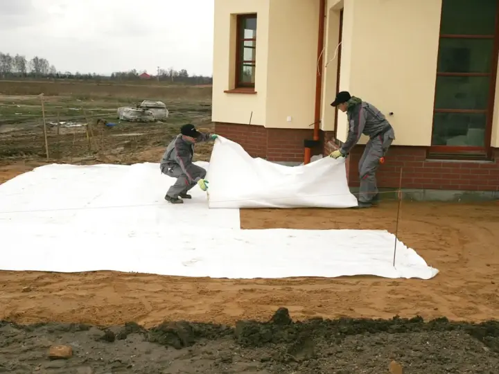 Dwóch robotników rozkłada białą geowłókninę, przygotowując tani taras na gruncie.