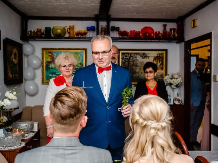 Uroczyste błogosławieństwo młodej pary przez starszego mężczyznę w niebieskiej marynarce.