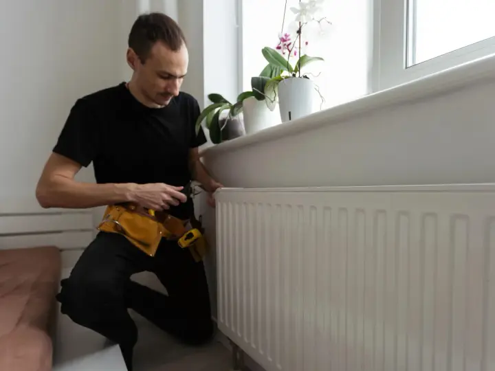 A man in a tool belt inspects a white radiator, checking for any signs of a cracked radiator before winter sets in.