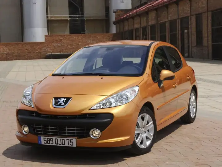 Pomarańczowy Peugeot 207 1.4 benzyna zaparkowany na brukowanym placu przed ceglanym budynkiem.