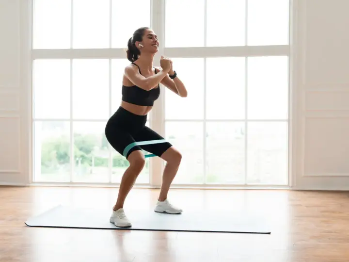 Femme souriante faisant un squat avec un élastique autour des cuisses, sur un tapis de yoga, devant une grande fenêtre lumineuse.