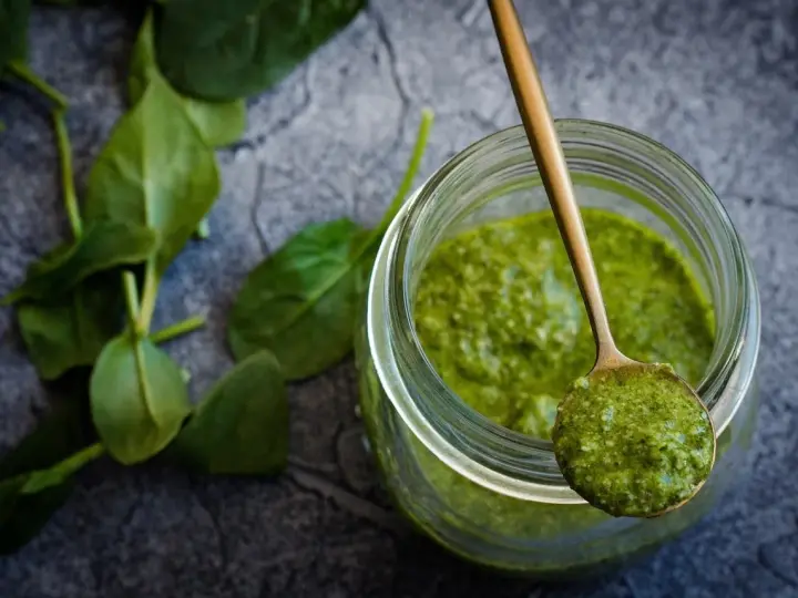 Słoik z zielonym pesto, na łyżce porcja sosu. Czy pesto ze słoika się podgrzewa? Świeże liście bazylii obok.