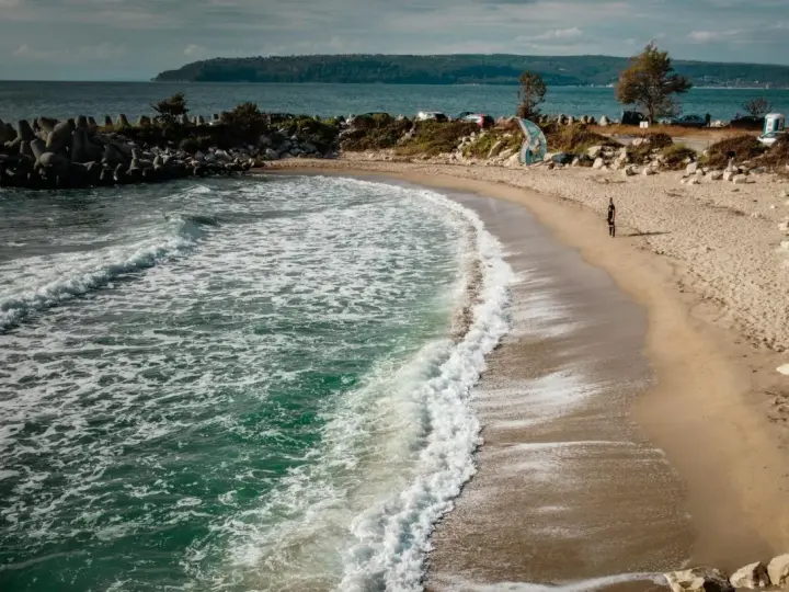 Piękne, turkusowe morze w Bułgarii obmywa piaszczystą plażę. W oddali widać wzgórza i samochody.