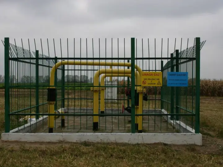 Żółty gazociąg wysokiego ciśnienia na działce, otoczony zielonym ogrodzeniem. Tablice ostrzegają przed gazem i ogniem.