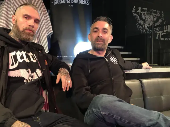 Deux hommes, dont un possible chanteur de Mass Hysteria, posent sur un canapé. L'un porte un sweat à capuche noir avec un aigle.