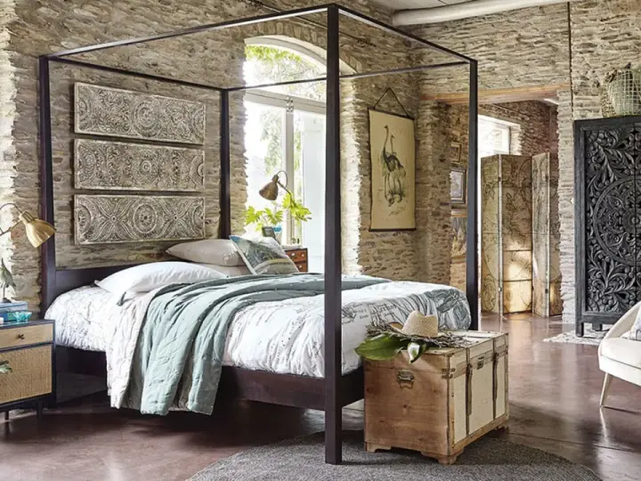 Ein Schlafzimmer mit Steinwänden und einem Himmelbett. Drei Wandpaneele mit orientalischen Mustern hängen über dem Bett. Ein Holztruhe steht am Fußende.