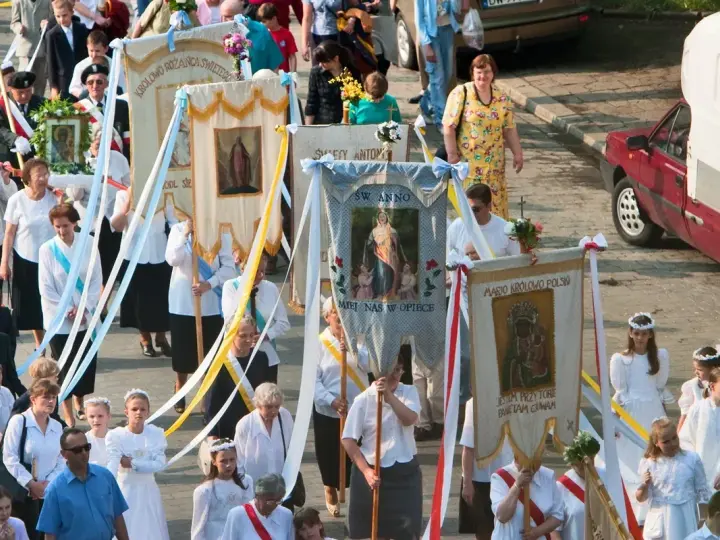 Procesja z banerami religijnymi, w tym z napisem "Boże Ciało 26". Ludzie niosą obrazy i kwiaty.