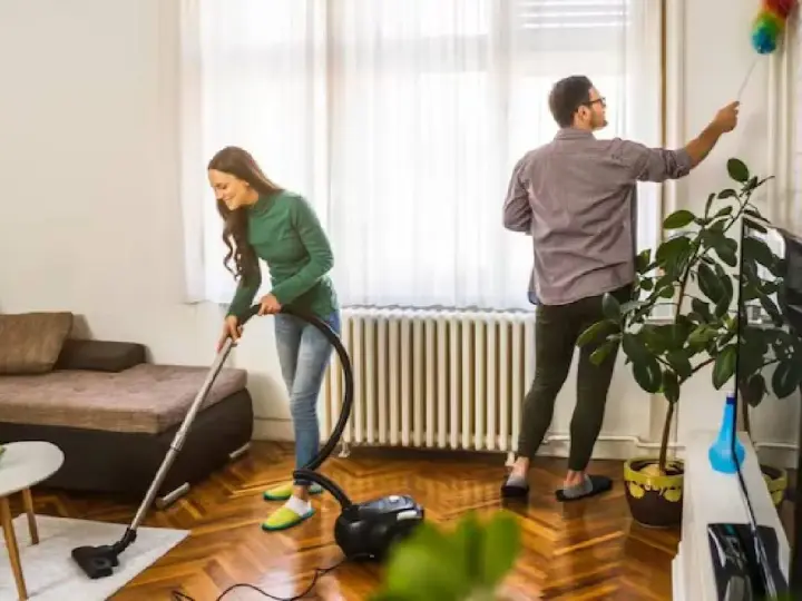 Pareja colaborando para purificar casa. Ella aspira el suelo, él quita el polvo de las paredes.