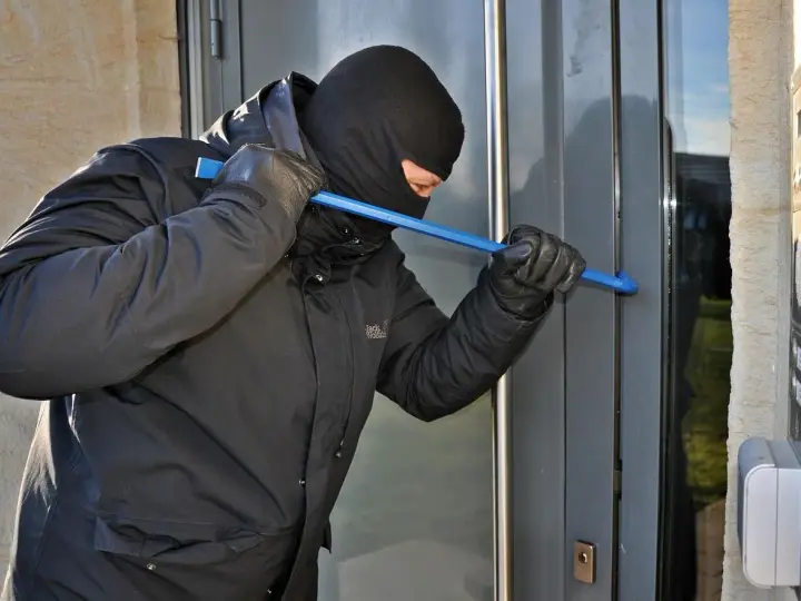 Osoba w kominiarce i rękawiczkach próbuje wyważyć drzwi łomem. To przykład kradzieży z włamaniem.
