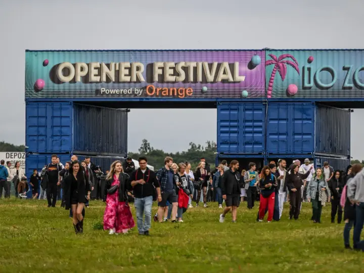 Tłum ludzi wchodzi na teren festiwalu. Nad nimi brama z napisem "OPEN'ER FESTIVAL". Kto będzie następny?