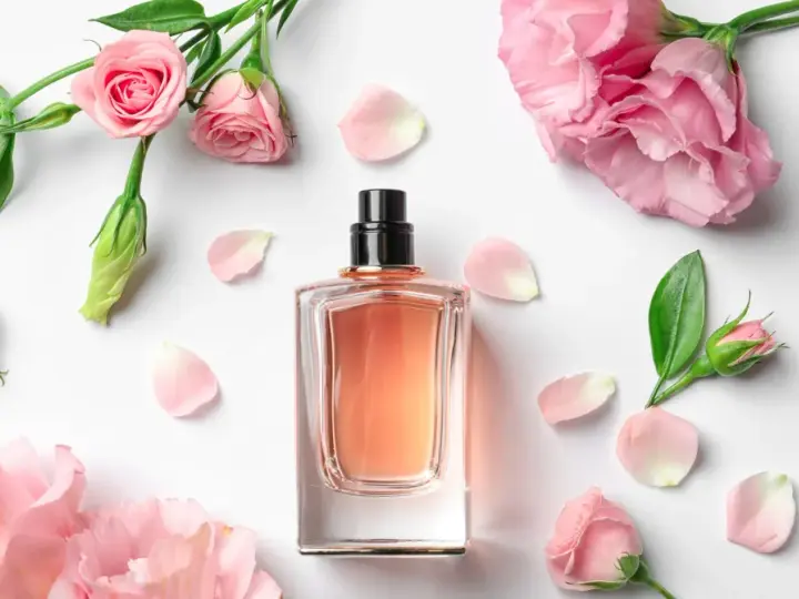 Elegancka butelka perfum z delikatnym, różowym płynem, otoczona pąkami i płatkami róż. Zapach jak słodka jeżyna.