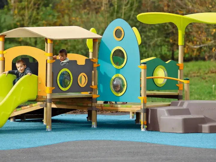 Des enfants jouent sur une aire de jeux colorée, respectant la réglementation aire de jeux extérieur. Un toboggan vert vif et une structure inspirée d'une fusée bleue.