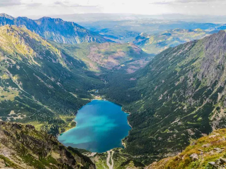 Widok na Morskie Oko z góry. W dole błękitna tafla jeziora otoczona lasem i górami. Kamera uchwyciła parking w oddali.