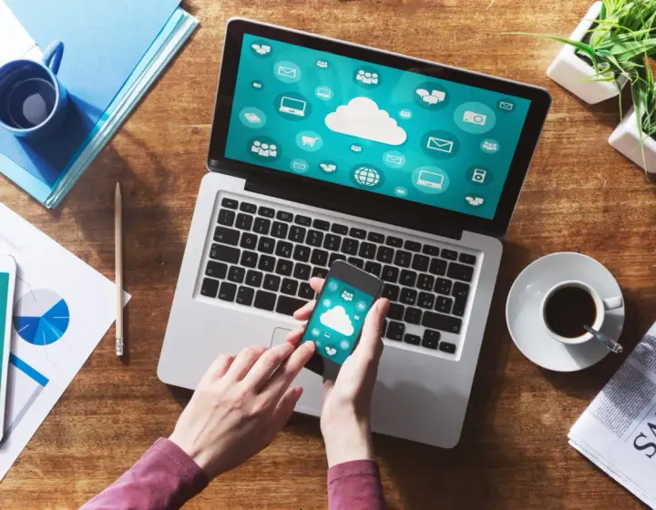 Dłonie trzymające smartfon z ikonami chmury na tle laptopa i dokumentów.